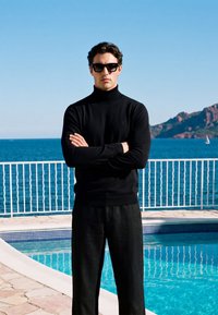 Homme habillé tout en noir avec des lunettes de soleil, debout les bras croisés près d'une piscine surplombant la mer bleue et la côte rocheuse sous un ciel clair.