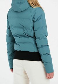 Türkisfarbene Pufferjacke mit Kapuze, ausgestattet mit einem schwarzen gerippten Saum. Gequiltete Textur und Reißverschlussdetails. Rückansicht, kombiniert mit hellen Hosen.