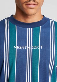 Modrá a tyrkysová pruhovaná tričko s bílými detaily. Na přední straně je vyšívaný nápis "NIGHTADDICT" bílou nití. Materiál z bavlněné směsi.