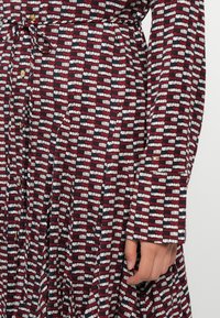 Sarkans, melns un balts rakstains kleita ar garām piedurknēm, piegulošu jostu un pogu detaļām. Audums šķiet gluds ar vieglu tekstūru.