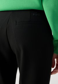 Pantaloni negri cu o croială ajustată, cu o textură fină, un buzunar posterior și un logo Lacoste discret, de culoare neagră, pe talie. O bluză verde este parțial vizibilă.