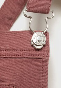 Overalls u ružičastoj boji od denima s metalnim detaljima, prilagodljivim trakama i šivenim detaljima. Tkanina ima glatku teksturu.