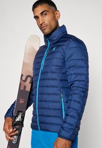 Veste isolante navy avec une fermeture éclair à l'avant, des accents bleus et un logo sur la poitrine. Le modèle tient un ski montrant ses fixations et son design.