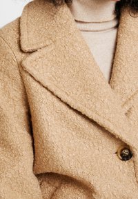 Beige textured coat with a notched lapel, crafted from boucle fabric, featuring a large button detail on the front.