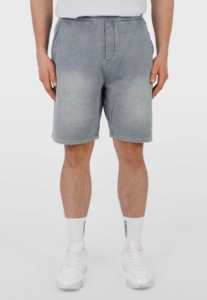 Mann trägt hellblaue Jeansshorts, weiße Sportsocken mit Logo, weiße Turnschuhe und ein weißes T-Shirt, steht mit den Händen an den Seiten.