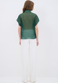 Blusa verde trasparente con un motivo testurizzato e maniche corte, abbinata a pantaloni bianchi a gamba larga. Il tessuto è leggero e fluido.
