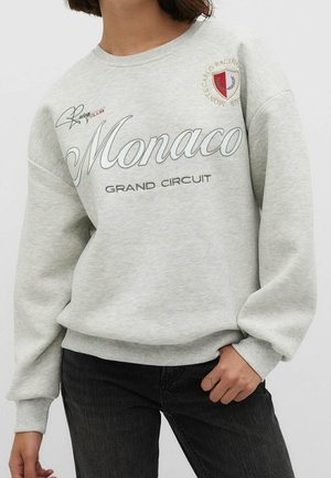 Femme portant un sweat-shirt gris clair avec le texte "Monaco Grand Circuit" et un écusson de club de course sur la poitrine, associé à un jean noir.