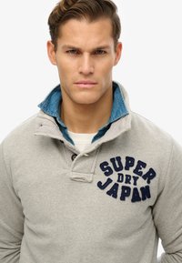 Szary polarowy pulowerek z zapięciem na guziki, z niebieską dżinsową wewnętrzną warstwą i granatowym haftem "SUPER DRY JAPAN."