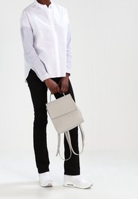 Bílá košile s dlouhým rukávem a pruhovanými rukávy, černé kalhoty, šedý obdélníkový batoh a bílé tenisky na osobě, která stojí.