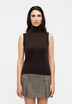 Donna con lunghi capelli scuri che indossa un maglione dolcevita senza maniche marrone scuro e una gonna plissettata grigia a quadri, in piedi davanti a uno sfondo bianco.