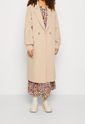 Vrouw draagt een beige lange jas over een kleurrijke bloemenjurk en lichtbeige enkellaarsjes, staand tegen een effen achtergrond.