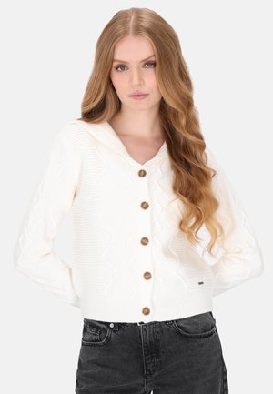 Ung kvinde med langt bølget hår iført en hvid kabelstrikket cardigan med knapper og sorte jeans, stående foran en ensfarvet baggrund.