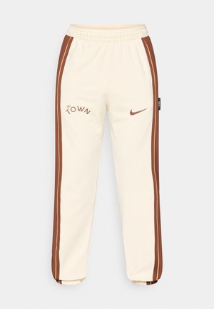 Cremefarbene Jogginghosen mit braunen Seitestreifen, die das Logo "The Town" und den Nike-Swoosh zeigen. Elastischer Bund und gerippte Bündchen. Weiches, glattes Material.