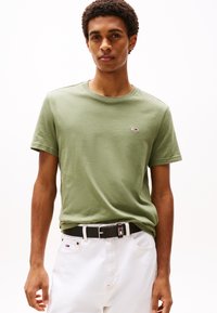 Tricou din bumbac de culoare verde măsliniu cu guler rotund, având un mic logo pe piept. Asortat cu pantaloni din denim albi și o curea neagră.