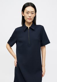 Vestido azul-marinho de manga curta com colarinho, apresentando um fecho éclair dourado na gola e um corte descontraído. Tecido liso e texturizado.