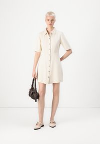 Robe crème à manches courtes avec un devant à boutons, un col décontracté et une silhouette droite. Le modèle tient un petit sac à main marron et porte des ballerines beiges.