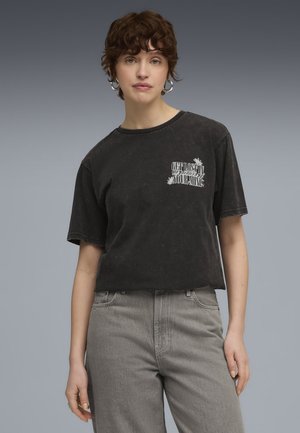 Schwarzes Baumwoll-T-Shirt mit entspannter Passform, kurzen Ärmeln und einem grafischen Druck auf der linken Brust. Kombiniert mit hellgrauen Jeans.
