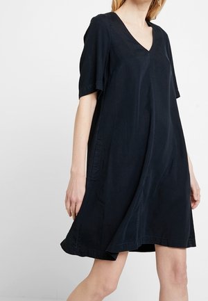 Frau in einem locker sitzenden, kurzärmeligen schwarzen Kleid mit V-Ausschnitt, die vor einem schlichten weißen Hintergrund steht.