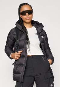 Casaco puffer preto com capuz, design acolchoado, fecho frontal e cordões ajustáveis. Logotipo branco no peito e um corte elegante.