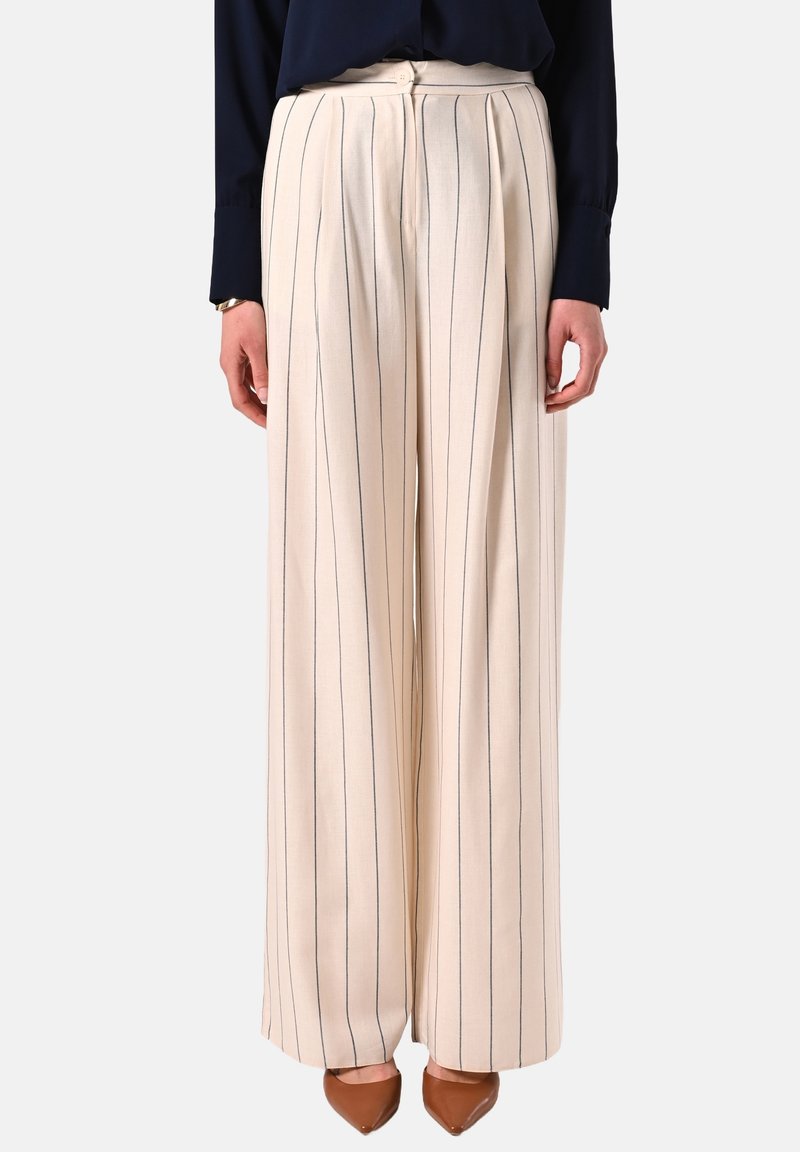 Femme portant un pantalon crème taille haute à jambes larges avec de fines rayures verticales, associé à des chaussures pointues marron et un haut à manches longues foncé.