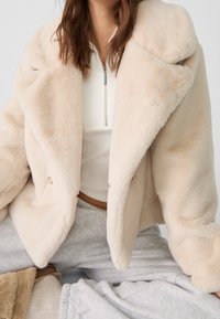 Beige zweireihige Jacke aus Kunstpelz mit übergroßem Kragen, weicher Struktur und zwei Knöpfen. Getragen über einem weißen, gerippten Oberteil und grauen Jogginghosen.