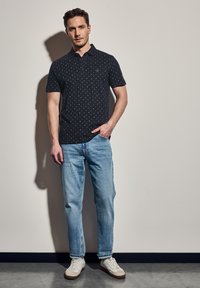 Černé polo tričko s jemným geometrickým vzorem, kombinované se svetle modrými džíny a bílými teniskami s béžovými akcenty.