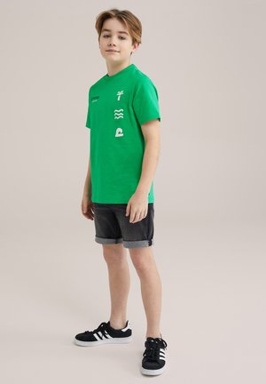 T-shirt verde in cotone con grafiche bianche, abbinata a pantaloni corti in denim scuro con orli arrotolati e sneakers nere con strisce bianche.