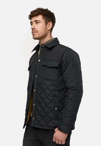 Quilted sort jakke med en ensfarvet sort overdel, specifikt frontlommer og trykknapper. Har en krave og er sat sammen med en lys halskrave.