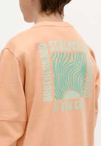 Brzoskwiniowy sweatshirt posiada duży projekt graficzny na plecach z napisem "BRATERSTWO" oraz falistymi liniami w kolorze turkusowym, a także motyw słońca.