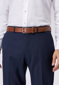 Bruine leren riem met een zilverkleurige gesp, gedragen over marineblauwe broeken. De riem heeft een gladde textuur en gaten voor aanpassing.