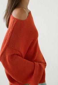 Strickpullover in Orange mit einem schulterfreien Design, lockeren Ärmeln und einer strukturierten Oberfläche. Vor einem neutralen Hintergrund platziert.