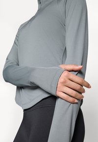 Person som har på seg en grå langermet idrettsgenser med tommelhull, krysser armene over svarte tettsittende leggins mot en hvit bakgrunn.