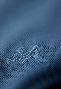 Niebieska tkanina o teksturowanym splocie, z wytłoczonym logo w prawym dolnym rogu. Projekt jest subtelny i monochromatyczny.