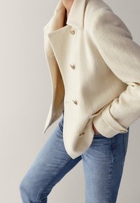 Giacca di colore crema con texture, colletto, design doppiopetto e bottoni a forma di cuore dorati, abbinata a jeans blu chiaro.