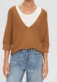 Brun stickad tröja med djup V-hals och 3/4 ärmar, lager på lager över en krämfärgad topp. Bärs med ljusblå denimjeans.