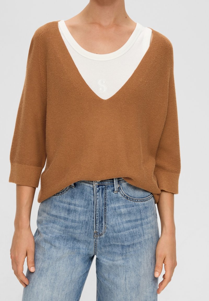 Brun stickad tröja med djup V-hals och 3/4 ärmar, lager på lager över en krämfärgad topp. Bärs med ljusblå denimjeans.