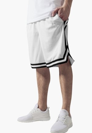 Person, die weiße Sportshorts mit schwarzem Rand und weiße Sportschuhe aus Netzmaterial trägt, steht mit einer Hand in der Tasche.
