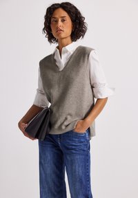 Kvinna i grå ärmlös tröja över vit skjorta och blå jeans, håller en svart handväska med ena handen i fickan.