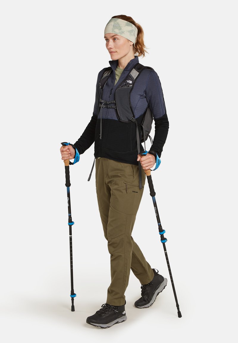 Schwarze und graue technische Weste über einem langärmligen Shirt, olivgrüne Hose, mit Trekkingstöcken, trägt ein leichtes Stirnband.