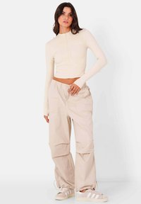 Beige cropped langemouwtop met een rits aan de voorkant, gecombineerd met loszittende beige cargo-broeken met grote zakken en elastische boorden, gedragen met sneakers.