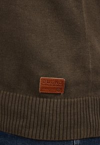 Olivekowy sweter z dzianiny z prążkowanym dołem, z brązową skórzaną metką, na której widnieje napis "BLEND GLOBAL JEANMAKER" na dole.