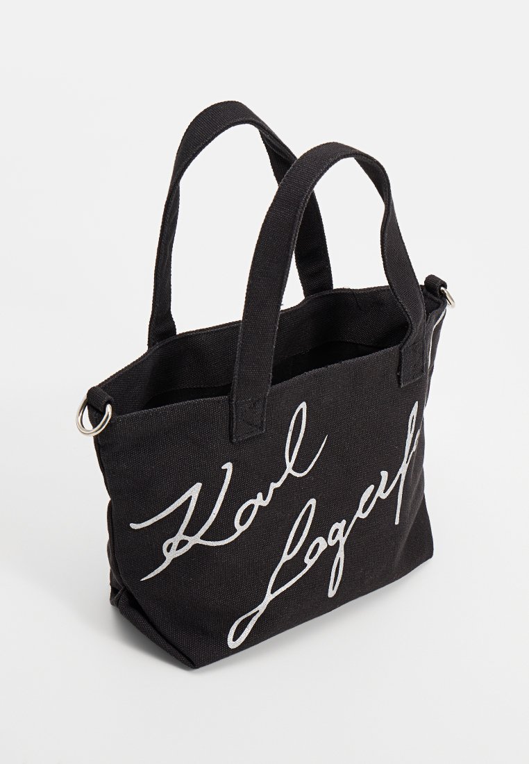 Sac fourre-tout en tissu noir avec deux anses et texte en cursive blanche "Karl Lagerfeld" sur le devant, détail en anneau métallique sur le côté.