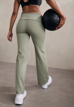 Person in hellgrüner Trainingshose und schwarzem Sport-BH hält einen schwarzen Medizinball hinter dem Rücken und steht auf grauem Boden.