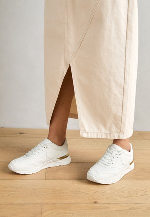 Person iført hvide sneakers med gyldne accenter og en beige nederdel med et slids bagpå, stående på et trægulv.