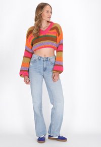 Maglione cropped lavorato a maglia con strisce audaci in arancione, rosa, verde e marrone; abbinato a jeans flare blu chiaro e sneaker viola.