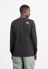 Schwarzes Langarmshirt aus Baumwolle mit rundem Ausschnitt und einem weißen Logo auf dem oberen Rücken. Trägt dazu hellgrüne Cargo-Hosen.