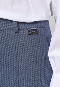 Pantalons bleus texturés à coupe slim, ornés d'une plaque en argent "HUGO" sur la ceinture, avec une blouse blanche visible au-dessus.