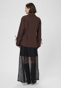 Woman wearing a brown long-sleeve blouse with tie details and a black lace maxi skirt with sheer panels, standing facing away on a plain background.