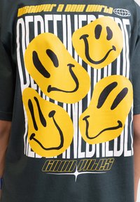 Grön bomulls t-shirt med stora gula smiley-ansiktsgrafik och fet text i vitt och gult. Mjuk textur, avslappnad design.