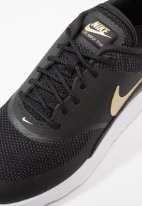 Black Nike Air Max shoe with gold accents, featuring a textured knit upper, sleek shape, and a white sole. Laces and logo prominently displayed.
